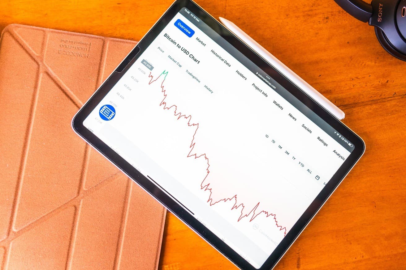 A tablet on an orange desk showing a Bitcoin-to-USD price chart, with a stylus beside it