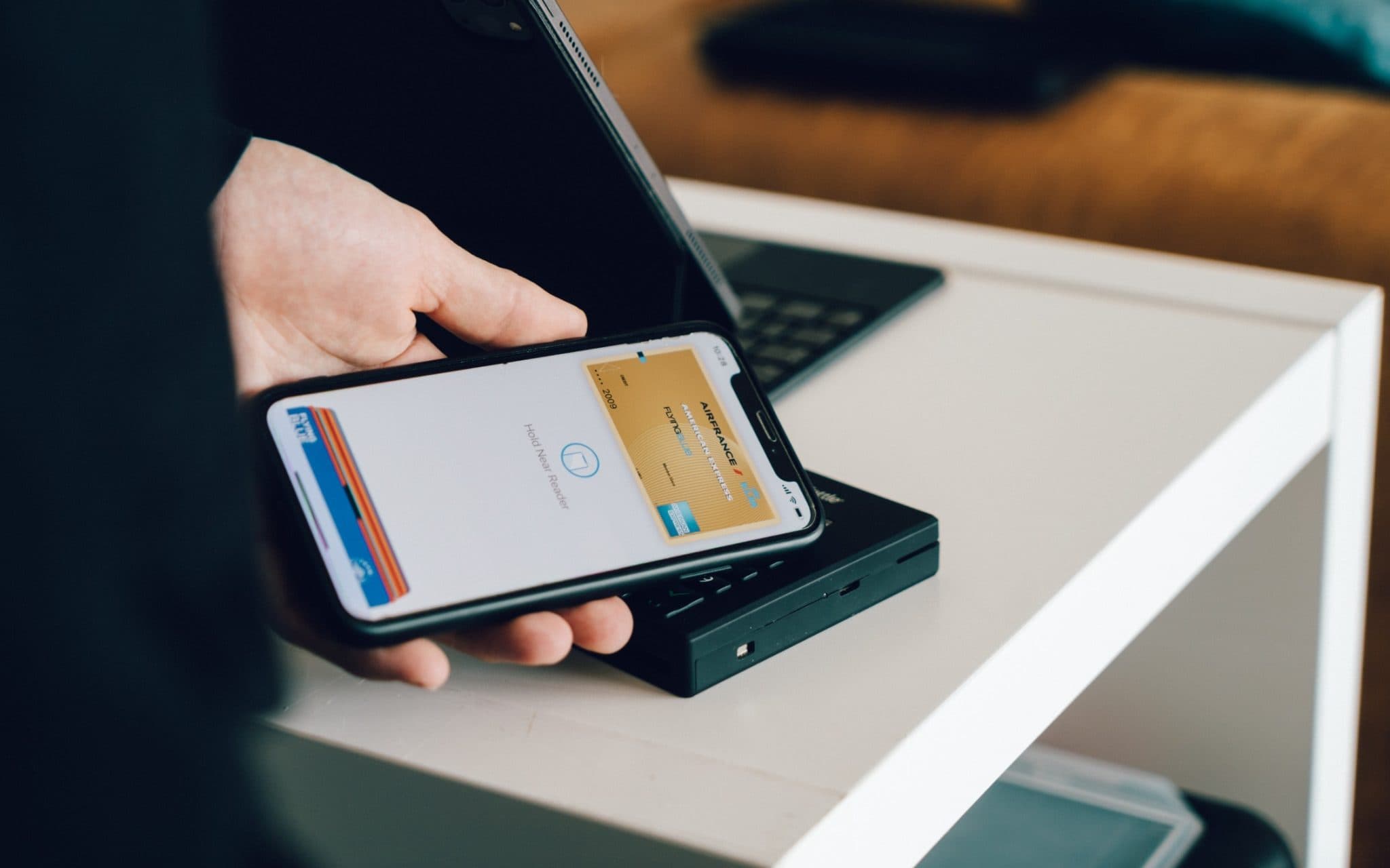 A hand holding a phone that shows a digital wallet with two payment cards, tapping a card reader on a desk